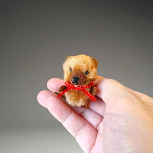 Op de afbeelding: Een miniatuur pluizig bruin berenfiguurtje met een donkerbruine snuit, kleine zwarte oogjes en een gestikte zwarte neus. De beer is versierd met een felrood lint gestrikt in een strik om de nek, met een zachte, textuurrijke vachtachtige uitstraling.