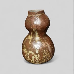 Puede incluir: Un jarrón de cerámica marrón con forma de calabaza. El jarrón tiene un cuello estrecho y una base más ancha, con una sección media bulbosa. La superficie es marrón moteado con vetas crema y beige. El jarrón es probablemente hecho a mano y tiene una estética rústica.