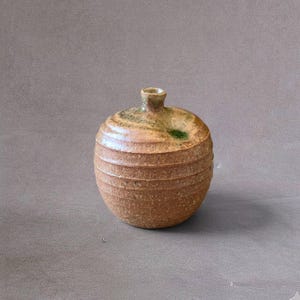 May include: A small, round, tan-colored ceramic vase with a textured, ribbed surface. The vessel features a narrow neck and a subtle, mottled green glaze accent near the top opening, showcasing a rustic, earthy appearance and a rounded base.