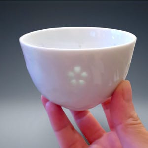 Puede incluir: Un cuenco de cerámica blanco con un diseño floral. El cuenco se sostiene en una mano y tiene un acabado liso y brillante. El diseño floral está formado por cinco pequeños recortes en forma de corazón.
