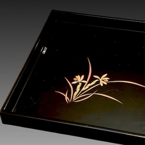 Puede incluir: Una bandeja rectangular de laca negra con un diseño incrustado en tono dorado de dos flores en flor con hojas largas y delgadas. La bandeja tiene un acabado liso y brillante, bordes elevados y un motivo botánico detallado.