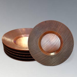 Puede incluir: Un conjunto de seis platos de madera con acabado cobrizo y un círculo central de madera. Los platos tienen una textura estriada en el borde exterior y un interior liso y claro. Los platos están dispuestos sobre una superficie gris claro.