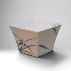 Puede incluir: Un cuenco cuadrado de cerámica blanca con base cónica y borde ensanchado. El exterior está decorado con un patrón botánico azul pintado a mano que consiste en hojas y tallos delgados que envuelven los lados del recipiente.