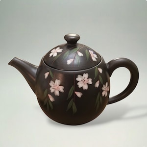 Puede incluir: Una tetera de cerámica marrón oscuro con cuerpo redondeado, boquilla y asa. La tetera está decorada con flores de cerezo blancas y rosas y hojas verdes. La tapa tiene un pequeño pomo.