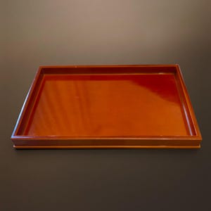 以下が含まれることがあります： 深みのある赤茶色の滑らかで光沢のある仕上げが施された長方形のトレイ。トレイは平らな底面と、周囲を囲むように立ち上がった真っ直ぐな縁が特徴で、シンプルで機能的なデザインを形成しています。