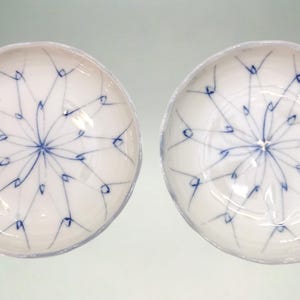 May include: Two white ceramic bowls with a blue floral design. The design radiates from the center of the bowl, resembling a starburst or dandelion. The bowls have a slightly irregular, wavy edge.