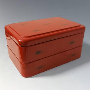 Op de afbeelding: Een rechthoekige, rode doos met twee niveaus en een glanzende afwerking. De doos heeft een deksel en is versierd met donkere, gevlekte accenten. Het ontwerp suggereert een opbergcontainer, mogelijk voor voedsel of kleine items.