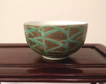 May include: A ceramic bowl with a brown exterior and a white interior. The bowl is decorated with a teal-colored bamboo leaf pattern. The bowl is sitting on a dark brown wooden table.