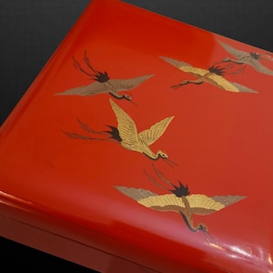 Puede incluir: Una caja lacada roja con un acabado brillante. La caja está decorada con grullas doradas y marrones en vuelo. Las grullas están representadas con las alas extendidas, creando una sensación de movimiento y elegancia. La caja tiene forma rectangular.