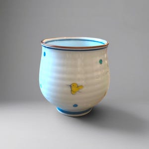 Può includere: Una tazza in ceramica bianca con corpo arrotondato e scanalato, caratterizzata da una piccola illustrazione di un uccellino giallo e pois sparsi color ottanio e blu. Il bordo è accentuato da una sottile linea marrone e la base ha due bande blu.