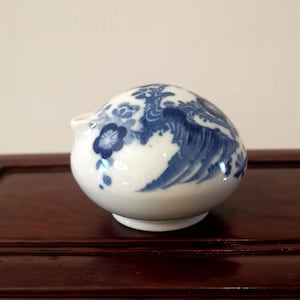 Puede incluir: Un pequeño recipiente de cerámica blanca con pico, con un diseño de olas y flores azules. El cuerpo redondo se asienta sobre una pequeña base. Probablemente utilizado para verter líquidos.