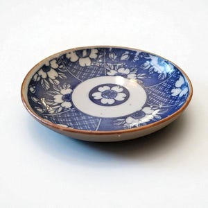 May include: A small, round ceramic dish with a blue and white floral pattern. The dish has a brown rim and a white center with a blue floral design. The blue pattern includes floral and geometric elements.