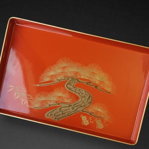 以下が含まれることがあります： 鮮やかな赤色の長方形の漆塗りサービングトレイ。トレイには、金色の、様式化された木のデザインが施されており、複雑なディテールが施されています。縁は細い金の縁でトリミングされており、この装飾品にエレガンスを添えています。