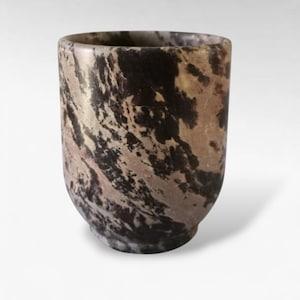 Puede incluir: Una taza cilíndrica de piedra con base redondeada. La taza presenta un patrón veteado de colores negro, marrón y beige. La taza es lisa y pulida, con un borde ligeramente ensanchado. La taza está vacía y se apoya sobre una superficie blanca.