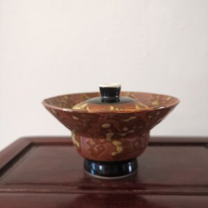 以下が含まれることがあります： 蓋付きの陶器のボウル。ボウルは茶色で、黄色と白のアクセントがあります。蓋は黒で、白と黒のノブが付いています。ボウルは黒いベースの上にあります。