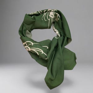 May include: A dark green scarf featuring a light-colored, thin-lined floral pattern. The fabric is draped in a loose, circular loop, showcasing the matte texture of the material and the contrast between the deep green background and the delicate lines.
