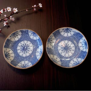 May include: Two blue and white ceramic plates with a floral design. The plates have a central flower motif surrounded by a geometric pattern. The rims are a light brown color. The plates are on a dark brown surface.
