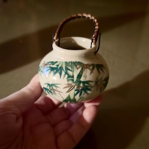Op de afbeelding: Een kleine, ronde keramische pot in crèmekleur met een craquelé glazuurafwerking. De buitenkant is voorzien van een geschilderd ontwerp van groene en bruine bamboestengels. Het heeft een gebogen, donkerbruin gevlochten handvat aan metalen lussen.