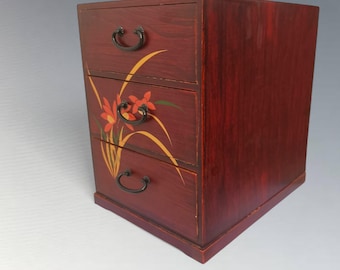 May include: A small, three-drawer wooden chest with a deep red finish. The drawers are adorned with a floral design in orange, yellow, and green. Each drawer has a black, curved handle. The chest is a compact, decorative storage piece.