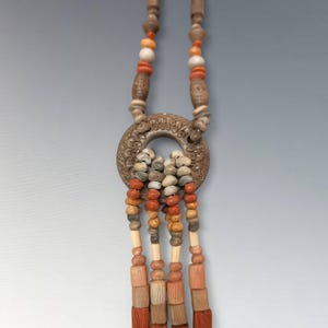 May include: A handcrafted necklace featuring a large, circular pendant with carved details and a cluster of small stones. The necklace has strands of wooden beads in shades of brown, orange, and beige, creating a unique, earthy aesthetic.
