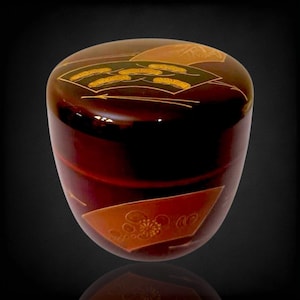 Puede incluir: Un recipiente japonés lacado, de color marrón oscuro, con tapa redondeada. La tapa y los lados presentan elementos decorativos dorados y marrones, incluyendo abanicos estilizados y motivos florales. El recipiente tiene un acabado brillante.