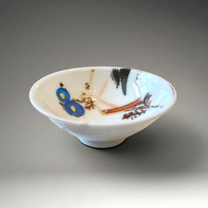 May include: A small, white ceramic bowl with a black base. The bowl is decorated with blue, brown, and black abstract designs. The bowl has a slightly flared rim and a smooth, glossy finish. The bowl is likely used for serving small portions of food.