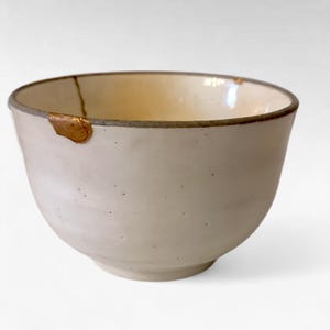 May include: A white ceramic bowl with a slightly irregular shape. The rim is a darker shade of gray, and a section of the bowl has been repaired with gold-colored material. The bowl has a minimalist aesthetic.