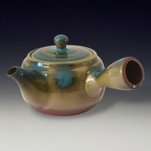 May include: A ceramic teapot with a side handle and a lid. The teapot is glazed in shades of green, brown, and blue, with a red base. The spout is on the left, and the handle is on the right. The lid has a small knob.