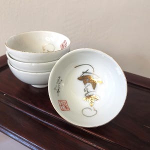 Puede incluir: Un juego de cuatro tazas de té de cerámica blanca con diseños pintados en oro y rojo. Las tazas están apiladas una encima de la otra y presentan un diseño de hoja de oro con caracteres japoneses.