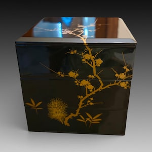 May include: A square, three-tiered black lacquer box featuring a glossy finish. The exterior is decorated with gold-colored illustrations of a flowering plum blossom branch, pine needles, and bamboo leaves that extend across the lid and sides.