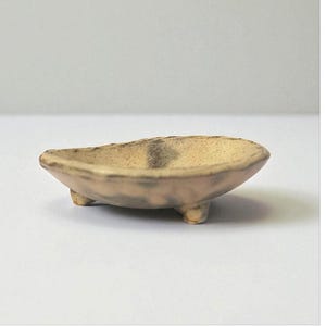 Pode incluir: Um pequeno prato de cerâmica feito à mão, de cor bege claro com uma faixa cinza escuro no centro. O prato, de formato oval, repousa sobre três pequenos pés arredondados. A estética geral é rústica e minimalista.
