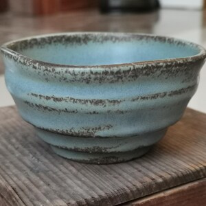 May include: A small, handmade ceramic bowl with a light blue glaze and a textured, ridged design. The rim and ridges have a darker, speckled finish. The bowl sits on a wooden surface, showcasing its rustic aesthetic.