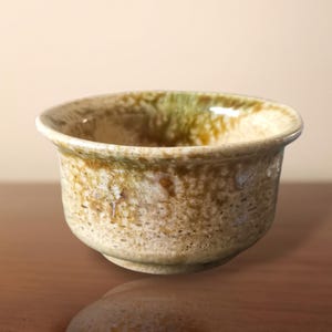 Könnte beinhalten: Eine kleine, handgefertigte Keramikschale mit strukturierter Oberfläche. Die Schale hat eine abgerundete Form mit einem leicht ausgestellten Rand. Die Farbpalette umfasst Erdtöne von Beige, Braun und Grün, wodurch eine natürliche, rustikale Ästhetik entsteht.