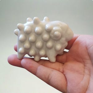 以下が含まれることがあります： 丸みを帯びた隆起のあるテクスチャ加工された表面を持つ、小さな白いセラミック製の羊の置物。 羊には4本の小さな脚と丸みを帯びた体があります。 置物は手で持たれており、そのサイズと詳細を示しています。