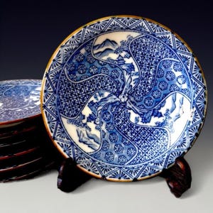 May include: Blue and white ceramic plate with a gold rim. The plate features a detailed landscape design with mountains and geometric patterns. The plate is displayed on a wooden stand, with a stack of similar plates in the background.