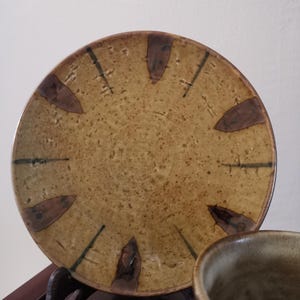 Può includere: Un piatto in ceramica fatto a mano e una ciotola abbinata. Il piatto è rotondo con uno sfondo marrone, dettagli marroni e linee verde scuro. La ciotola è di un colore e consistenza simili. Il piatto ha un diametro di circa 20 cm.