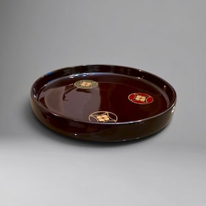 Könnte beinhalten: Ein rundes, dunkelbraun lackiertes Tablett mit glänzender Oberfläche. Auf der Innenseite befinden sich drei kreisförmige Motive, die jeweils eine stilisierte vierblättrige Blume in Gold-, Rot- und gedeckten Grüntönen zeigen.