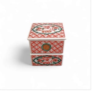 Puede incluir: Pequeña caja de cerámica de dos niveles con un patrón de entramado rojo y blanco. La caja presenta diseños florales decorativos verdes y rojos y un círculo naranja central. Probablemente para guardar objetos pequeños.