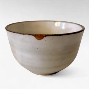 May include: A white ceramic bowl with a slightly irregular shape. The rim is dark brown, and a small area of gold-colored repair is visible. The bowl has a minimalist aesthetic, suitable for serving food or as a decorative piece.