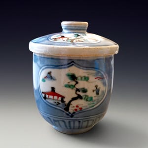 Könnte beinhalten: Eine blau-weiße Keramik-Teetasse mit Deckel. Die Tasse zeigt eine handgemalte Landschaftsszene mit einem roten Gebäude, einem Baum und Figuren. Der Deckel hat ein ähnliches Design. Die Tasse ist etwa 10 cm hoch.