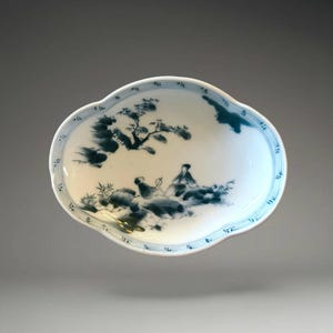 May include: An oval ceramic dish with a scalloped edge, featuring a white base and blue hand-painted ink-wash style illustrations of a tree, rocks, and two figures sitting outdoors. The rim is decorated with thin blue concentric line patterns.