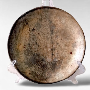 May include: A handmade, round ceramic plate with a textured surface. The plate has a light brown center with darker brown edges and a speckled appearance. It is displayed on a clear plastic stand.