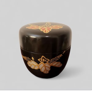 Puede incluir: Un recipiente negro y redondo con tapa, con diseños florales dorados y rojos. El recipiente tiene un acabado brillante y un patrón floral alrededor de la base. La tapa también tiene un diseño floral.