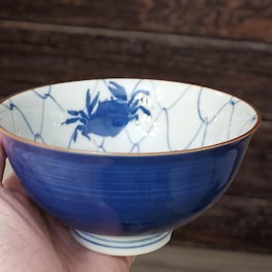 Puede incluir: Un cuenco de cerámica azul y blanco con un diseño de cangrejo. El cuenco presenta una ilustración de cangrejo azul en el interior, con un patrón de red. El exterior es de color azul liso y el borde tiene un ribete dorado. El cuenco se sostiene en una mano.