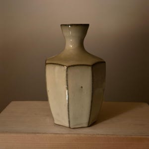 Könnte beinhalten: Eine sechseckige Keramikvase mit cremefarbener Glasur und einem schmalen Hals. Die Vase weist dunkle, definierte Kanten entlang ihrer sechs vertikalen Seiten auf, was eine strukturierte geometrische Form mit einer glatten, glänzenden Oberfläche erzeugt.