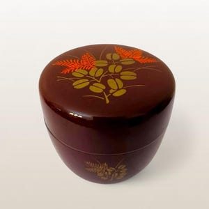 Puede incluir: Un recipiente redondo, marrón oscuro, con un acabado brillante. La tapa está decorada con un diseño floral dorado y naranja. El recipiente tiene un diseño floral a juego en la base. El recipiente probablemente se utiliza para el almacenamiento.