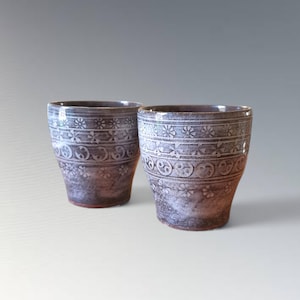 Op de afbeelding: Twee keramische bekers met een donkere, gevlekte afwerking. Elke beker heeft een herhalend patroon van bloemen en cirkelvormige ontwerpen in een lichtere kleur, waardoor een textuur ontstaat. De bekers hebben een licht uitlopende vorm.