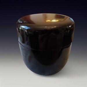Puede incluir: Un recipiente negro brillante con forma ovalada y redondeada. El recipiente tiene tapa y una superficie lisa y reflectante. El color oscuro y el diseño sencillo sugieren un artículo de almacenamiento, posiblemente para cosméticos o objetos pequeños.