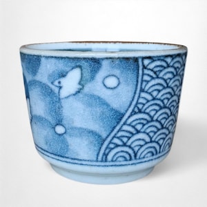 May include: A small, blue and white ceramic cup with a rounded base. The cup features a bird and wave pattern design. The rim is a darker blue, and the cup has a smooth, glossy finish. The cup is approximately 7.6 cm in diameter.