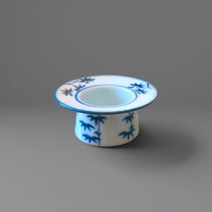 May include: A white ceramic egg cup featuring a wide, flat rim and a cylindrical base. The exterior and the top rim are decorated with hand-painted blue botanical motifs, including delicate leaf and bamboo-like patterns, with a blue trim.